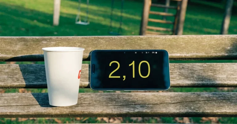 Ein Smartphone mit dunklem Display liegt auf einer Parkbank, daneben ein Kaffeebecher, im Hintergrund ein Spielplatz.